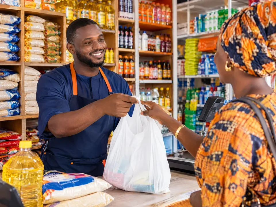 Un boutiquier tend un sac de courses à une cliente, devant ses rayons de riz, d’huile et de boissons